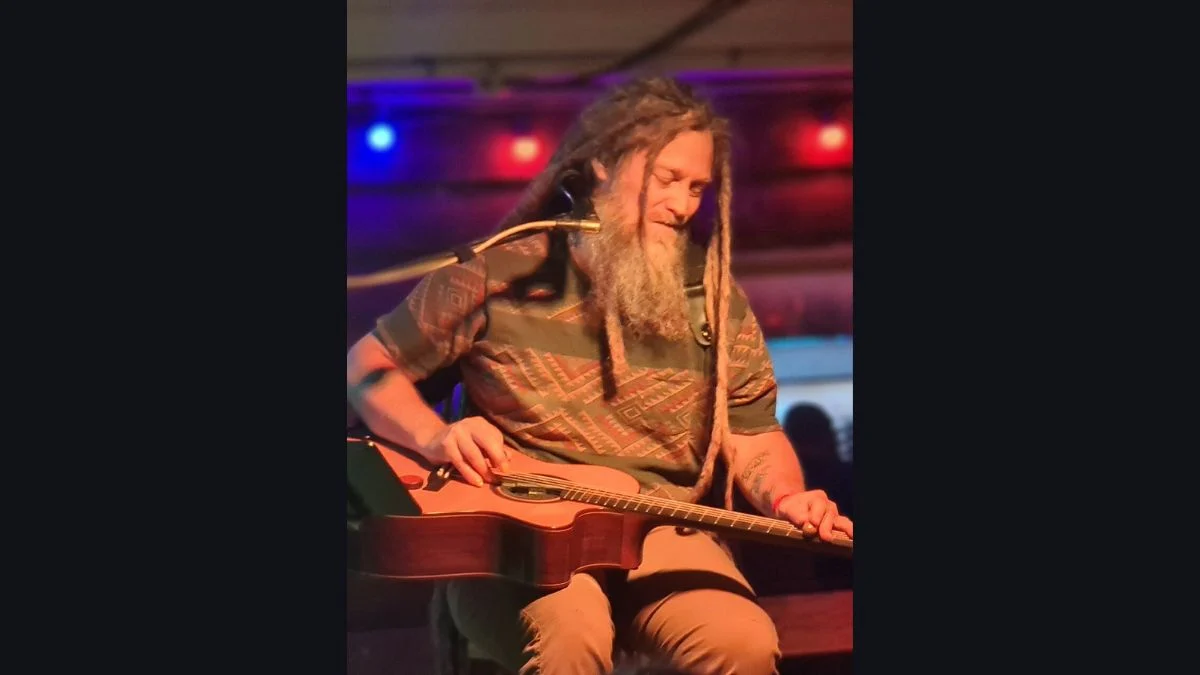 A musician with long dreadlocks playing an acoustic guitar on a brightly lit stage.