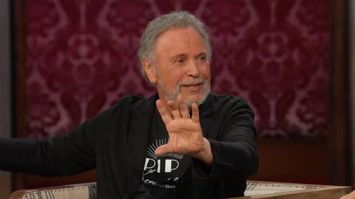Billy Crystal speaking animatedly during an interview while raising his hand.