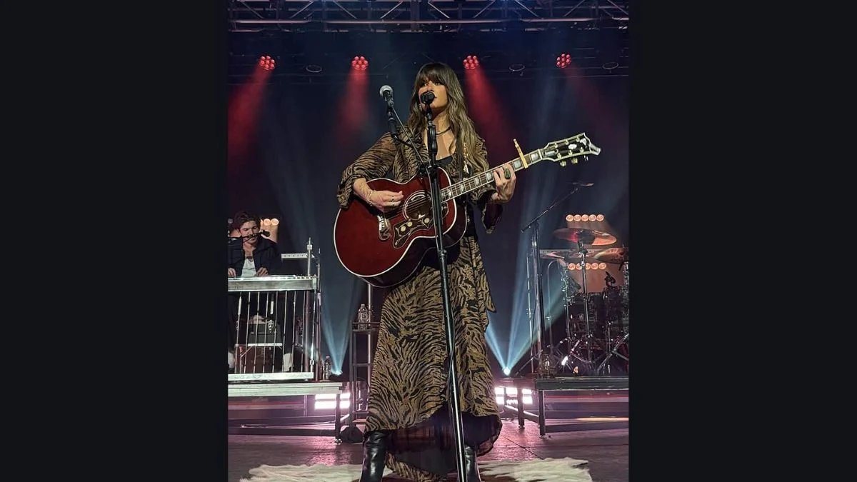 Ella Langley wearing a brown patterned outfit with a guitar on stage under concert lights.