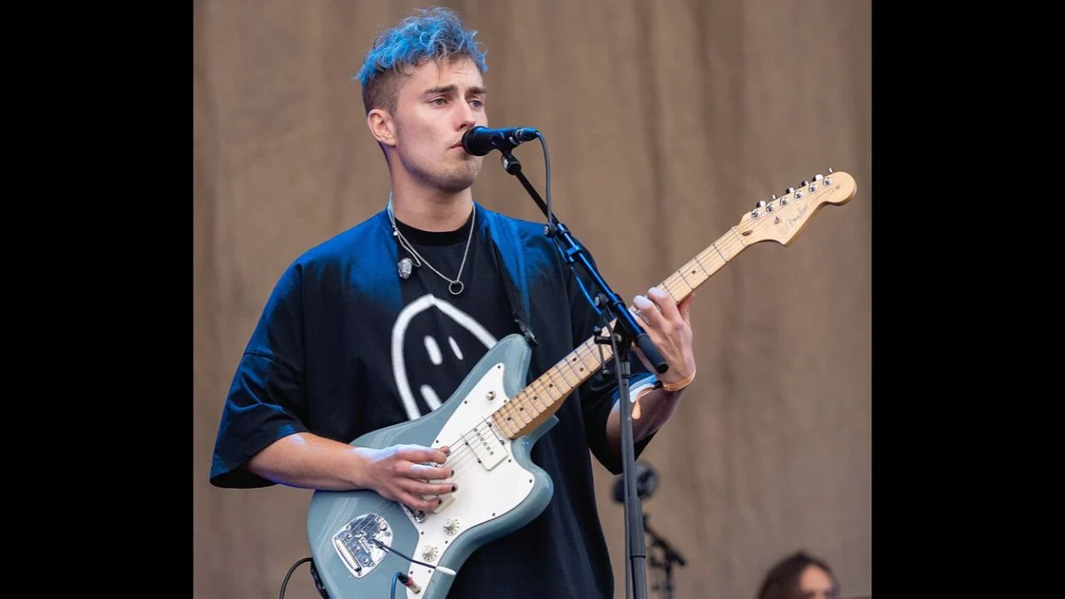 Sam Fender playing a blue electric guitar and singing into a microphone on stage