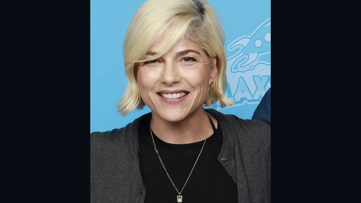Actress Selma Blair smiling with blonde hair and a grey cardigan in front of a blue backdrop.