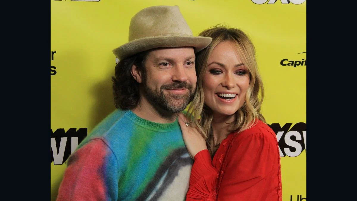 Jason Sudeikis wearing a hat and tie-dye sweater posing alongside Olivia Wilde in a red top on a yellow backdrop.