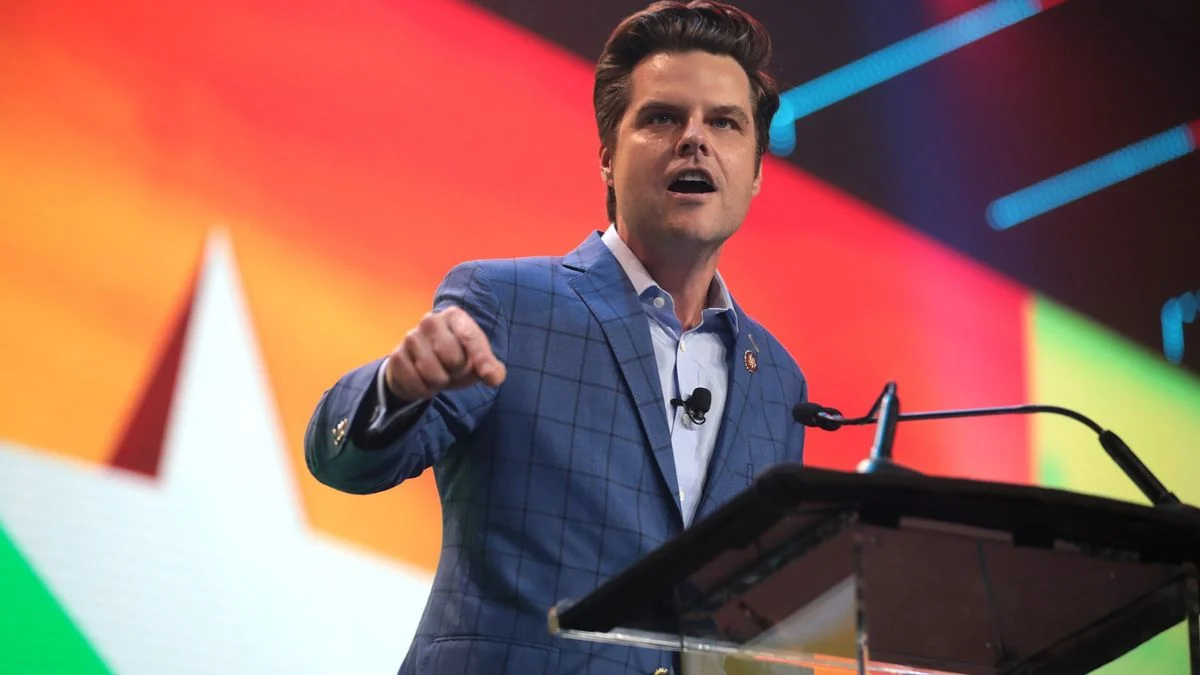 Matt Gaetz speaking at a podium while wearing a blue plaid suit jacket against a colorful background.