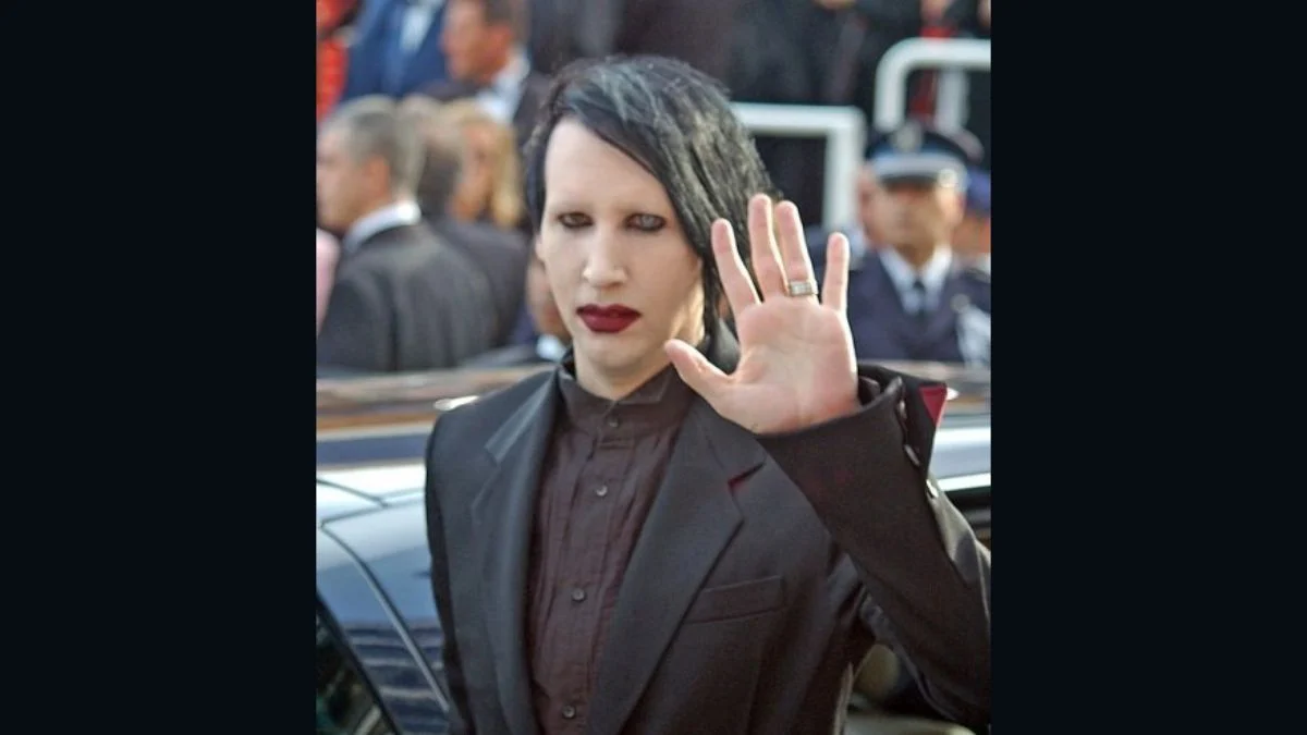 Musician Marilyn Manson wearing a black suit and dark makeup raising his open hand in a wave.