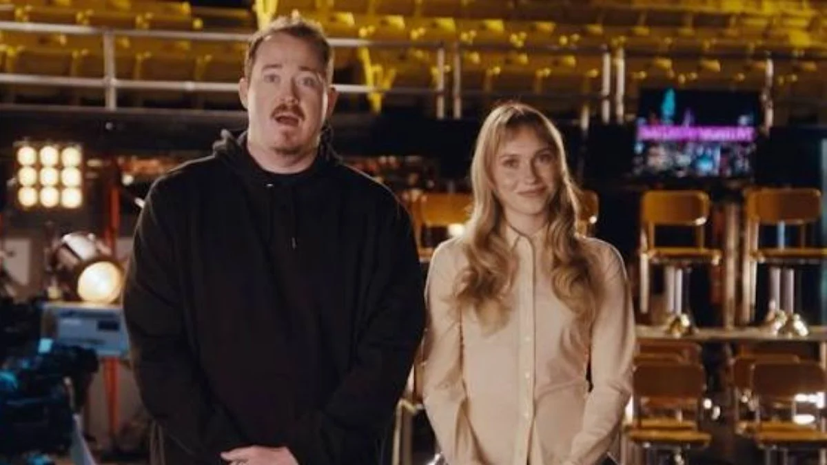 Comedian Shane Gillis and blonde cast member standing together on the Studio 8H set filming a promo for Saturday Night Live.