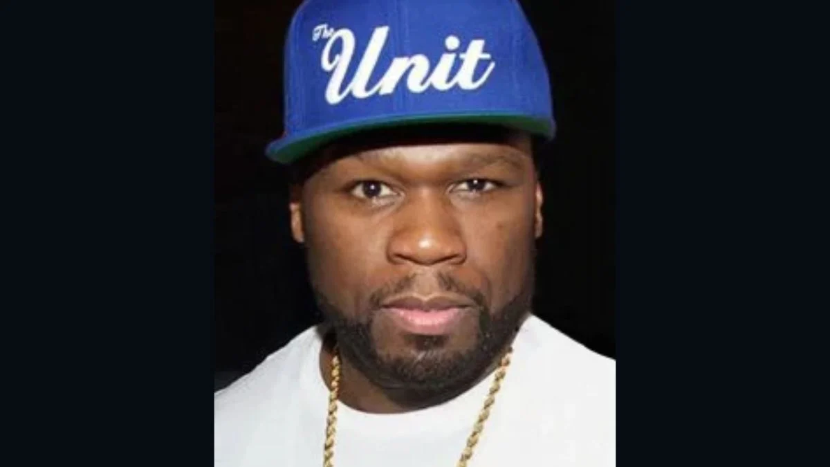50 Cent wearing a blue baseball cap reading "G Unit" and a gold chain looking forward