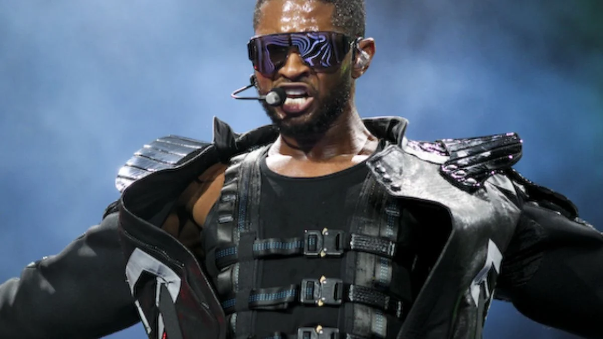 Usher performing live on stage wearing futuristic sunglasses, a headpiece microphone, and an embellished tactical-style black outfit.