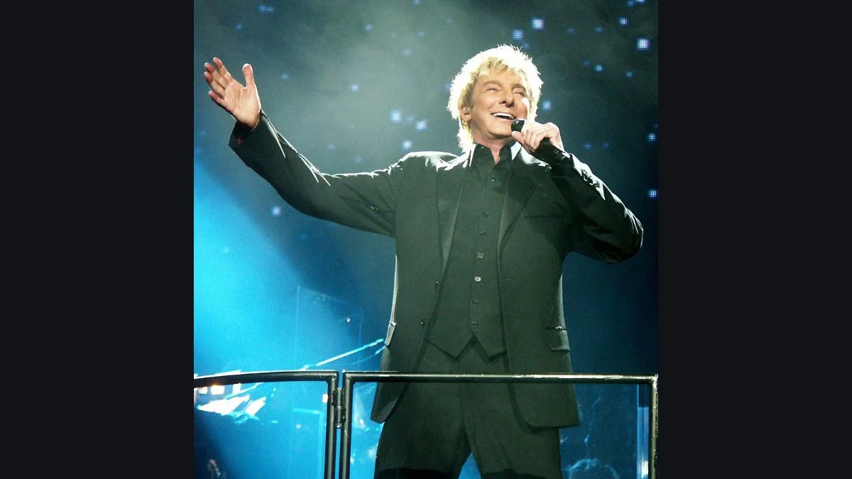 Barry Manilow performing live on stage with a microphone under spotlight.