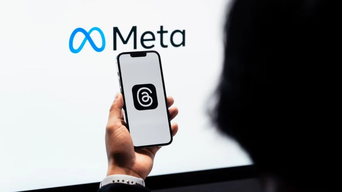 Person holding a smartphone displaying the Meta Threads logo in front of a screen with the blue Meta company logo.