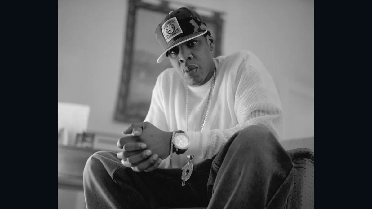 Black and white portrait of rapper Jay-Z sitting with hands clasped wearing a cap and chain