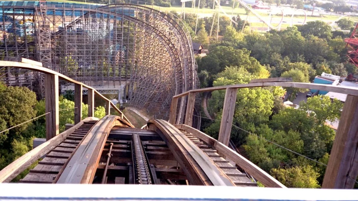 First-person view looking down the wooden track drop of a roller coaster surrounded by trees.