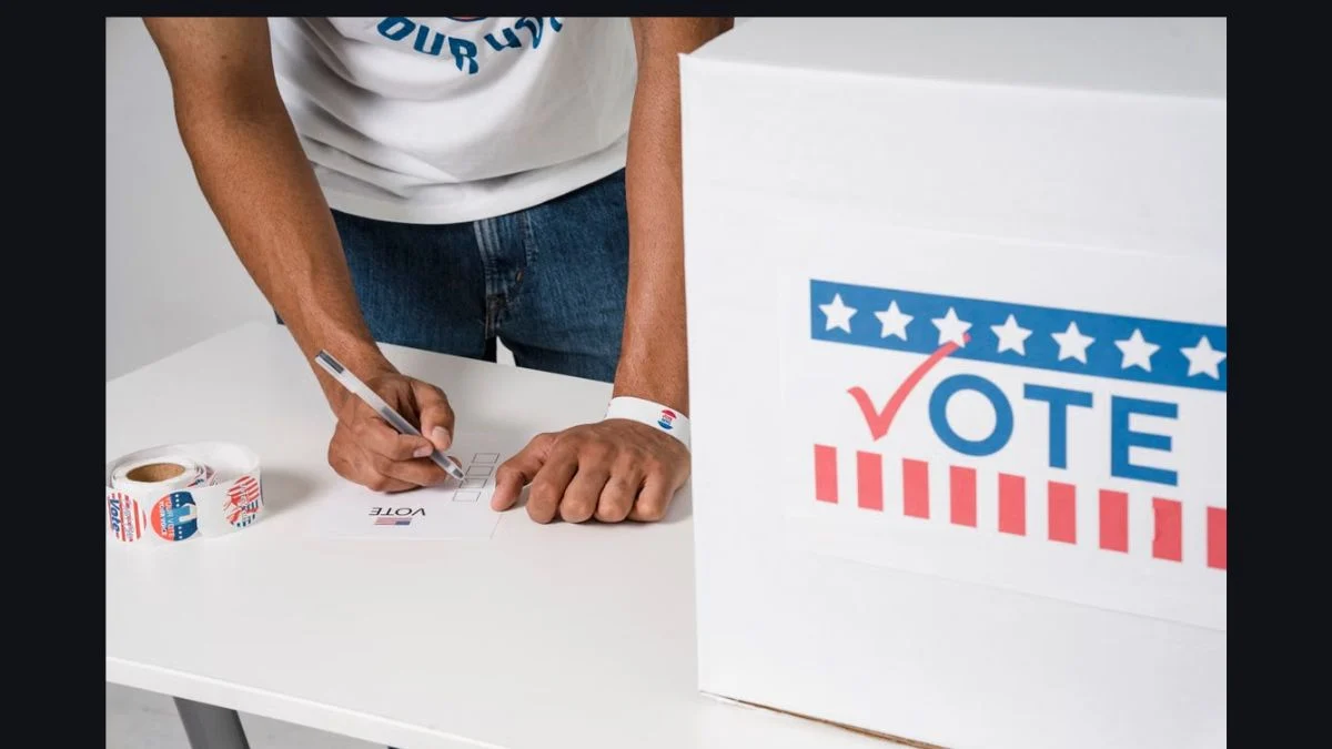 A person casting a ballot into a white box labeled VOTE with stars and stripes.