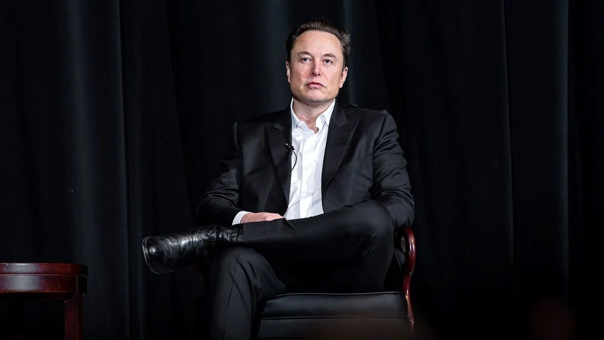 Tech billionaire Elon Musk seated in a dark suit jacket and boots during an event on stage.