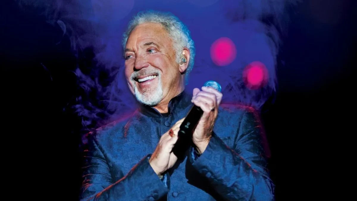 Sir Tom Jones smiling and holding a microphone on stage surrounded by blue stage lighting