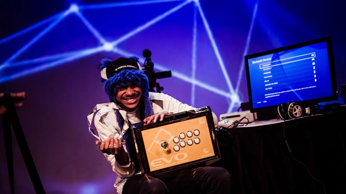 Fighting game champion SonicFox smiling in a blue fuzzy hat holding an EVO trophy and arcade fight stick
