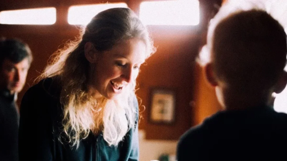 Elizabeth Holmes smiling as she leans down to speak with a child indoors with a man visible in the background.