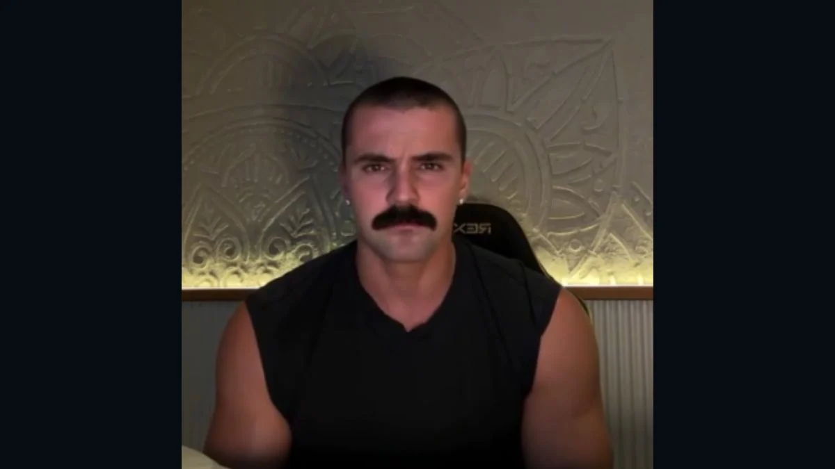 British fitness influencer George Armstrong speaking in a video with a mustache and black tank top.