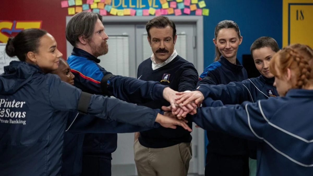 Jason Sudeikis as Ted Lasso and Brendan Hunt as Coach Beard joining hands in a huddle with members of the AFC Richmond women's football team.
